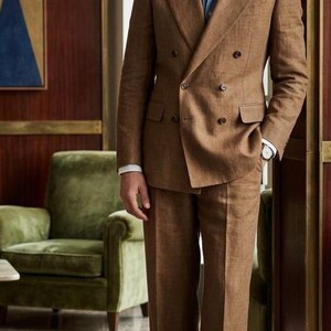 May include: A brown double-breasted suit jacket and matching trousers worn with a light blue button-down shirt. The suit features a textured fabric, wide lapels, and dark buttons. The trousers have a straight-leg cut with a cuffed hem.