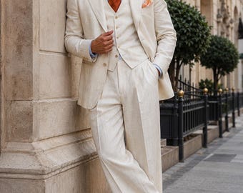 May include: A three-piece cream-colored suit featuring a blazer, a button-up vest, and pleated trousers. The ensemble is paired with a light blue collared shirt, a solid burnt orange necktie, a matching pocket square, and two-tone brown and white shoes.