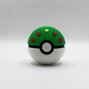 Op de afbeelding: Een bolvormig object met een groene bovenhelft en een witte onderhelft, gescheiden door een zwarte band. Het groene gedeelte bevat vier kleine rode driehoekige accenten en een enkele gele ronde knop in het midden van de bovenkant.