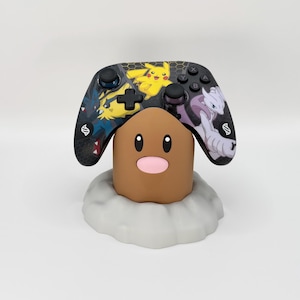 Pode incluir: Um suporte cilíndrico castanho com nariz cor-de-rosa e olhos pretos sobre uma base rochosa cinzenta. Um comando de jogo sem fios preto repousa no topo, decorado com ilustrações coloridas de Pikachu, Lucario e Mewtwo, com botões pretos.
