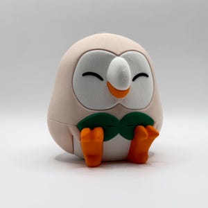 May include: A 3D printed figurine of a round, light pink owl-like creature with a white facial disc, closed eyes, and an orange beak. It features a green bowtie-like chest detail and orange feet, all rendered with a visible layered texture.
