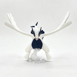 Peut inclure: Une figurine de Lugia blanche et bleu foncé avec des ailes déployées, une tête pointue et une queue, positionnée au-dessus d'une base texturée translucide imitant l'apparence d'éclaboussures d'eau, sur un fond blanc uni.