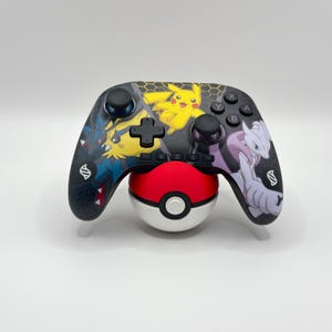 Pode incluir: Um comando de videojogos sem fios preto com uma impressão gráfica colorida de Pikachu, Lucario e Mewtwo. O comando inclui sticks analógicos pretos, um pad direcional e botões de ação, apoiado num suporte esférico vermelho e branco.