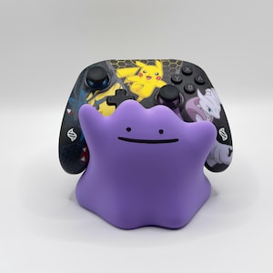 Könnte beinhalten: Ein violetter, klecksförmiger Controller-Ständer mit einem einfachen Gesicht, bestehend aus zwei Punkten als Augen und einem Strich als Mund. Dahinter befindet sich ein schwarzer kabelloser Game-Controller mit bunten Pikachu- und Mewtu-Motiven.
