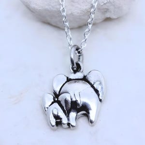 May include: A silver-toned metal necklace featuring a pendant shaped like two elephants, one larger and one smaller, connected by a jump ring to a delicate silver chain. The pendant has a polished finish with dark recessed areas to define the shapes.