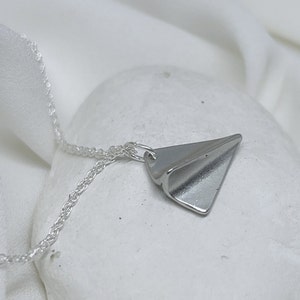 May include: A silver necklace with a folded paper airplane pendant. The necklace is on a white surface.
