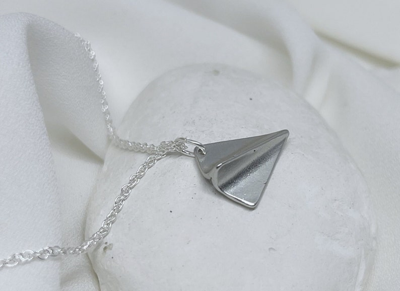 May include: A silver necklace with a folded paper airplane pendant. The necklace is on a white surface.