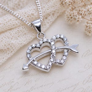 May include: A silver-toned necklace featuring a pendant with two interlocking heart shapes. The hearts are outlined with small, clear, sparkling stones and are pierced by a single silver arrow that extends across the centre of both heart forms.