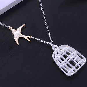 Könnte beinhalten: Eine silberne Kettenkette mit einem silbernen Vogelkäfig-Anhänger und einem goldenen Vogel-Anhänger. Der Vogel fliegt aus dem Käfig.