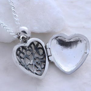 Puede incluir: Un collar con un dije de coraz&oacute;n de plata con un dise&ntilde;o intrincado en la parte delantera y un interior liso y vac&iacute;o. El dije est&aacute; abierto, revelando el interior.