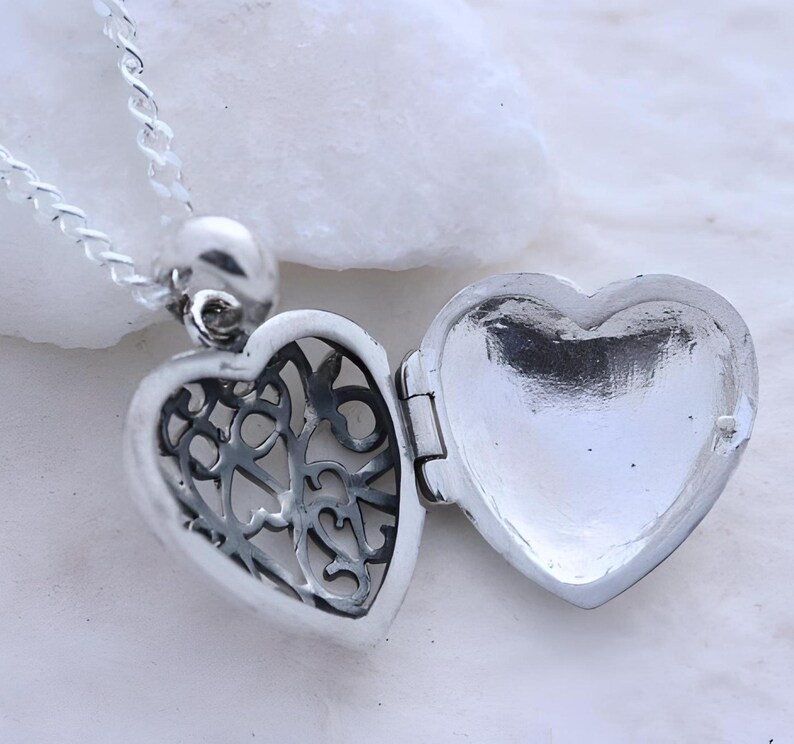 Puede incluir: Un collar con un dije de coraz&oacute;n de plata con un dise&ntilde;o intrincado en la parte delantera y un interior liso y vac&iacute;o. El dije est&aacute; abierto, revelando el interior.