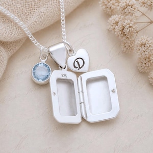 May include: A sterling silver locket necklace featuring a rectangular open locket stamped with 925, a small heart charm engraved with the letter D, and a round light blue gemstone charm suspended from a delicate silver chain.
