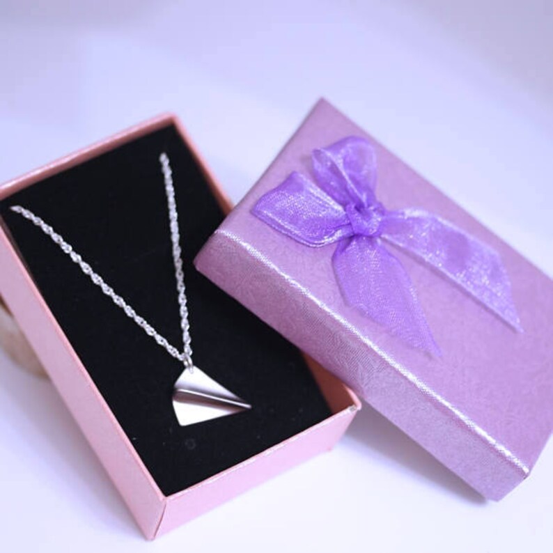 May include: A silver paper airplane necklace in a pink gift box with a purple ribbon.