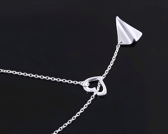 May include: A silver-toned necklace featuring a delicate chain, a small open heart-shaped charm, and a polished paper airplane pendant. The metal components have a smooth, reflective finish, creating a clean and simple design for this jewelry piece.