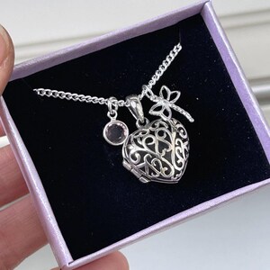 Puede incluir: Un collar de plata con un dije en forma de coraz&oacute;n, un dije de lib&eacute;lula y un peque&ntilde;o cristal rosa. El collar se muestra en una caja de terciopelo morado.