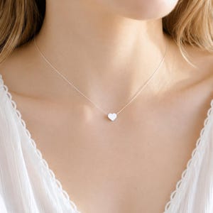 May include: A delicate silver-toned necklace featuring a small, smooth, solid heart-shaped pendant centered on a thin, minimalist chain. The metal has a polished, reflective finish that catches the light, creating a simple and clean aesthetic.