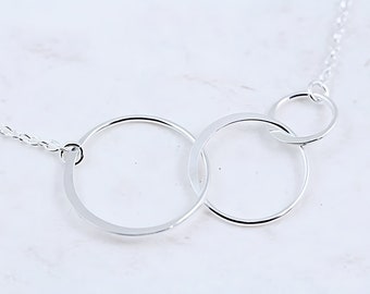 Pode incluir: Um colar de prata com três anéis circulares entrelaçados de tamanhos variados. Os anéis de metal polido estão ligados a uma delicada corrente de prata, criando um design de elos contínuos disposto horizontalmente.