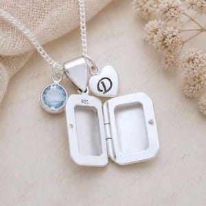 May include: A silver locket necklace with a chain, a blue gemstone charm, and a heart-shaped charm with the letter "D". The locket is open, revealing a space for a photo. The necklace is on a light-colored surface.
