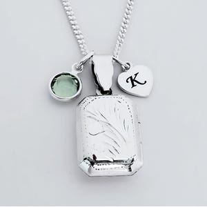 Op de afbeelding: Een zilveren ketting met een rechthoekig medaillon, een groene edelsteen en een hartvormige bedel met de letter "K". Het medaillon heeft een gegraveerd bloemmotief. De ketting is een gourmette.