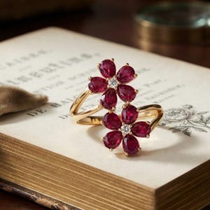 May include: A gold bypass ring featuring two flower-shaped motifs, each composed of five pear-cut deep red rubies surrounding a single small round white diamond centre. The band is crafted from polished yellow gold, creating a floral design.