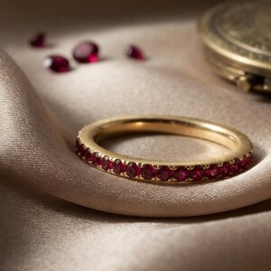 Könnte beinhalten: Ein goldener Eternity-Ring mit einer durchgehenden Reihe kleiner, runder, tiefroter Rubine, die in das Metall eingefasst sind. Der Ring liegt auf einem glatten, drapierten Stoff, wobei einige lose rote Rubine daneben liegen.