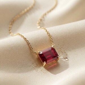 May include: A gold chain necklace featuring a rectangular emerald-cut deep red gemstone pendant held in a four-prong setting. The pendant is suspended from a delicate gold link chain, showcasing the vibrant red color of the stone.