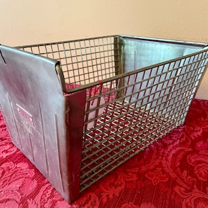 May include: A silver metal wire basket with a solid metal front panel. The basket has a rectangular shape and is sitting on a red patterned surface.