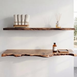 Peut inclure: Deux étagères murales horizontales en bois naturel avec des bords bruts. L'étagère supérieure porte un support en fil doré avec quatre serviettes blanches pliées et un diffuseur à roseaux. L'étagère inférieure porte un flacon en verre brun.