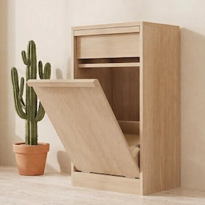 May include: A light oak wood cabinet featuring a top drawer and a large, tilt-out front panel designed for waste or storage. The cabinet has a clean, rectangular frame with a visible wood grain texture and a smooth, light brown finish.