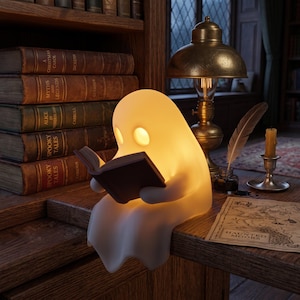 May include: A glowing, off-white, ghost-shaped lamp sits on a wooden desk, holding a small open book. The lamp features two circular, illuminated eye cutouts. It is positioned near a brass desk lamp, an inkwell with a quill, and a map.