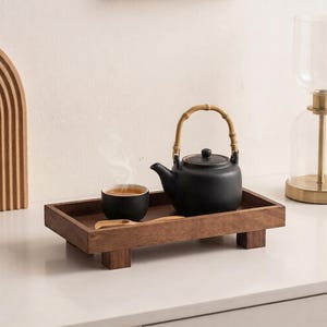 Könnte beinhalten: Eine mattschwarze Keramik-Teekanne mit gebogenem Bambusgriff und eine passende schwarze Keramiktasse mit Tee, beide auf einem rechteckigen Holztablett mit vier kurzen Beinen, ergänzt durch einen kleinen Holzlöffel neben der Tasse.