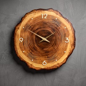 Peut inclure: Une horloge murale ronde fabriquée à partir d'une tranche de bois naturel présentant des cernes visibles dans des tons marron et beige. Le cadran comprend des aiguilles en métal doré et des chiffres dorés aux positions 12, 3, 6 et 9 heures, avec de petits points dorés marquant les autres heures.