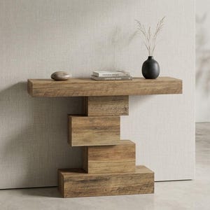 Puede incluir: Una mesa consola de madera con una superficie rectangular y una base formada por cuatro bloques rectangulares apilados. Sobre la mesa hay una piedra gris lisa, dos libros apilados con texto en el lomo y un jarrón negro con tallos de hierba.