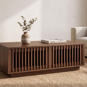 Puede incluir: Una mesa de centro rectangular de madera oscura con dos puertas frontales que presentan detalles de listones verticales. La superficie de la mesa sostiene un pequeño jarrón de cerámica con tallos de eucalipto y un libro con la palabra tome impresa.