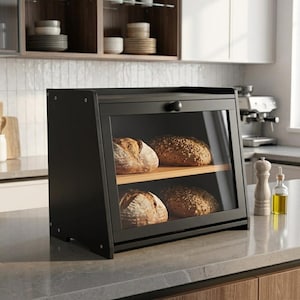 May include: A black wooden bread box with a clear glass front door and a round knob. The interior features two shelves, each holding a round loaf of crusty artisan bread. The unit has a slanted top design and sits on a flat countertop surface.