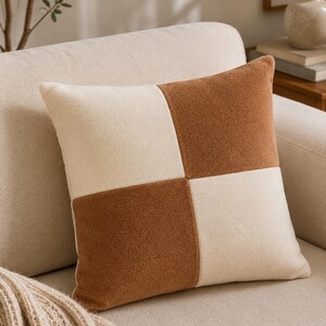 Peut inclure: Un coussin carré avec un motif en damier composé de blocs alternés de couleur crème et brun chaud. Le tissu présente une finition douce, texturée et semblable à de la polaire. Le coussin est formé de quatre quadrants distincts, créant un design géométrique équilibré.