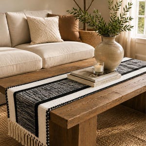 Pode incluir: Um caminho de mesa retangular com um padrão tecido de riscas pretas e brancas com um acabamento texturado. O caminho de mesa é rematado com longas franjas de cor creme ao longo das extremidades curtas, criando uma borda decorativa.
