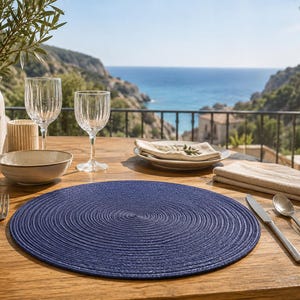 Peut inclure: Un set de table rond tissé bleu marine avec un motif en spirale concentrique, posé sur une table en bois avec une fourchette, un couteau et une cuillère en argent, un bol en céramique, deux verres côtelés, une assiette et un vase.