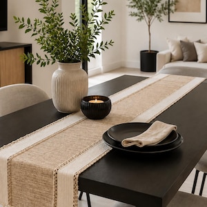 Peut inclure: Un chemin de table rectangulaire composé d'un panneau central en tissu tissé beige, bordé de bandes crème avec des détails en crochet complexes et terminé par des pompons suspendus aux extrémités, posé sur une table à manger en bois sombre.