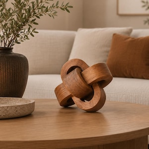 Può includere: Una scultura decorativa in legno composta da tre anelli circolari intrecciati con una finitura marrone naturale e liscia. L'oggetto presenta un design annodato continuo con venature del legno visibili, appoggiato su una superficie piana.