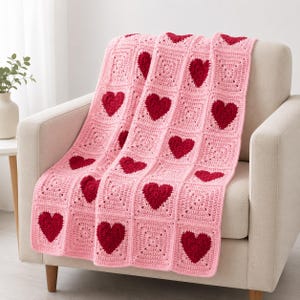 May include: A handmade crochet blanket featuring a repeating pattern of square blocks. Each block is crafted from light pink yarn and contains a centred, solid dark red heart motif. The blanket has a textured, grid-like appearance throughout.