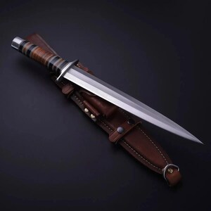 May include: A fixed-blade knife with a double-edged steel blade and a stacked leather handle featuring black and silver accents. The knife is accompanied by a brown leather sheath with visible stitching, metal rivets, and a metal D-ring attachment.