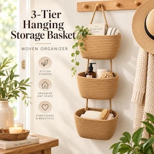 May include: A 3-tier hanging storage basket made of tan woven rope, suspended by thick cords from a wooden wall hook. The organizer features three oval-shaped baskets containing a small potted plant, a bottle, and folded white textiles.