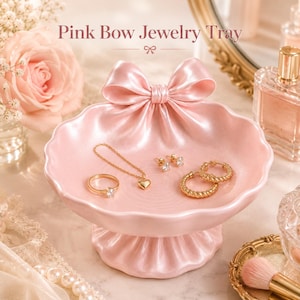 May include: A light pink jewelry tray featuring a sculpted bow at the top and a scalloped edge. The tray is pedestal-style with a pleated base. Inside the tray are gold-toned jewelry pieces including a ring, a heart pendant necklace, and earrings.