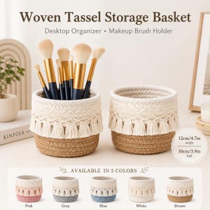 May include: Two round woven storage baskets with cream-coloured macrame-style trim and tassels over a natural brown jute base. One basket holds makeup brushes with gold ferrules. Text reads Woven Tassel Storage Basket, 12cm/4.7in wide, 10cm/3.9in tall.