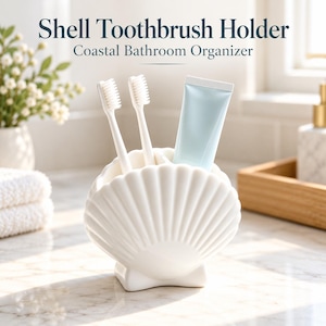 Puede incluir: Un soporte para cepillos de dientes de cerámica blanca con forma de concha marina y estrías verticales. Contiene dos cepillos de dientes blancos y un tubo de pasta dental azul claro. El texto Shell Toothbrush Holder Coastal Bathroom Organizer es visible.