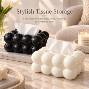 May include: Two rectangular tissue box covers with a bubble-like, spherical texture. One cover is glossy black and the other is glossy white. Each box has a central opening at the top with white tissues protruding from the center of the structure.