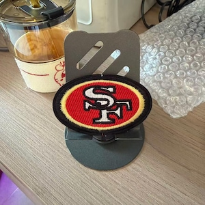 Puede incluir: Un parche bordado de forma ovalada que presenta el logotipo de los San Francisco 49ers con un monograma SF blanco sobre un fondo rojo, rodeado por un borde dorado y un borde exterior negro grueso, exhibido en un soporte de plástico gris oscuro.