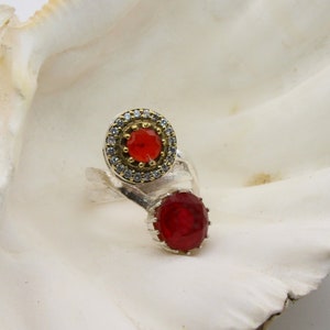 Könnte beinhalten: Ein silberner Ring mit zwei roten Edelsteinen. Der größere Edelstein ist von einem Heiligenschein aus kleineren weißen Edelsteinen umgeben. Der Ring hat ein einzigartiges, asymmetrisches Design.