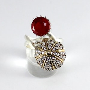 May include: A silver ring with a red gemstone and a round, white gemstone cluster. The ring is on a white display stand.